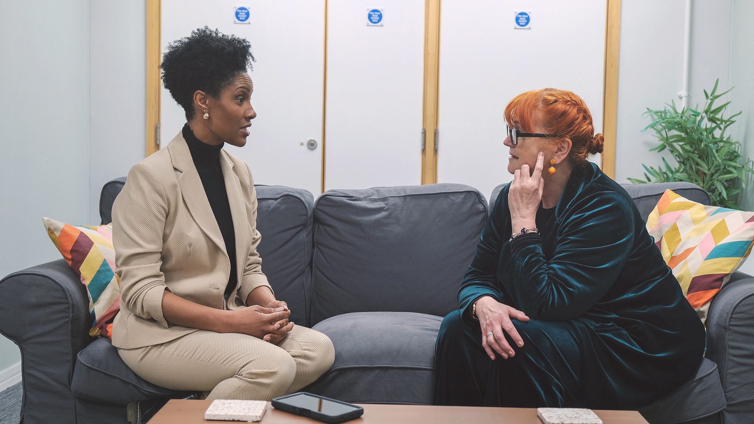 two people face eachother, having a conversation on a grey sofa. There is a light wood coffee table in front of them and a white wall in the background