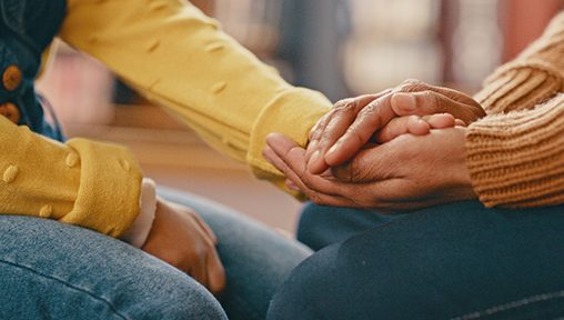 One person in a yellow top comforts another person in an orange jumper by holding their hand