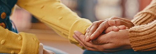 One person in a yellow top comforts another person in an orange jumper by holding their hand
