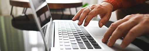 an image of a silver laptop, with two hands to the right of the image reaching toward the keyboard to type. the person is wearing an orange shirt.